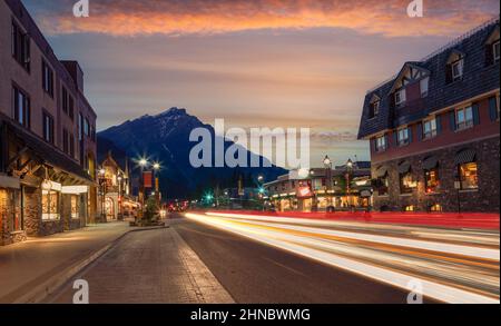 Sonnenuntergang auf der Banff Avenue im Banff National Park mit dem Cascade Mountain im Hintergrund, während leichte Wege von Autos die Straße während des Nachts erleuchteten. Stockfoto