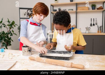 Süße blonde Junge rolling hausgemachter Teig auf Holztisch mit seiner Mutter in der Nähe von durch Während zusammen kochen Stockfoto