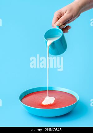 Frauenhand gießt Creme in die Tomatensuppe. Köstliche Tomatencremesuppe in einer Schüssel isoliert auf blauem Hintergrund Stockfoto