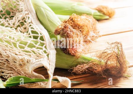 Vier süße frische grüne Milch Maiskolben Ohrblätter liegen in Öko wiederverwendbare Einkaufstasche auf Holztisch Hintergrund. Maisbraun lockiges Haar. Natur ökologisch Stockfoto
