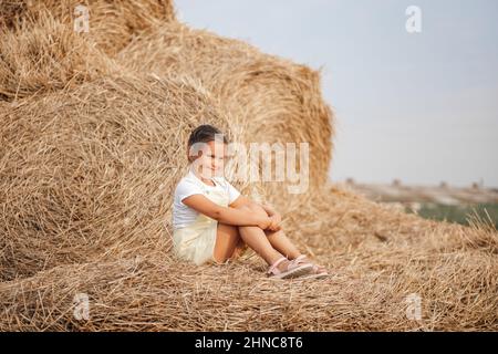 Kleines weibliches Kind mit listigen Gesichtsausdruck auf Heuhaufen, das irgendwo hin leicht lächelnd aussach. Zeit weg von der Stadt auf dem Land Feld mit Tonnen Stockfoto