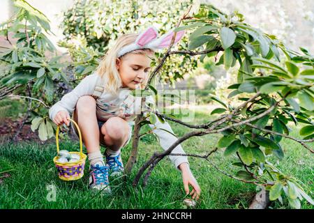 Liebenswert kleines Mädchen trägt Hasen Ohren hält einen Korb mit Ostereiern im Freien am Frühlingstag. Kind hat Spaß auf Ostereiersuche. Kinder suchen nach Stockfoto