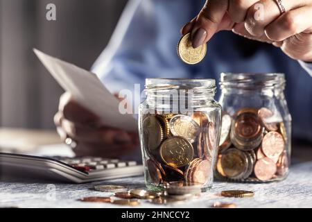 Eine Frau, die eine Papierrechnung in der Hand hält, fügt in ein Glas voller Euromünzen Cent ein. Stockfoto