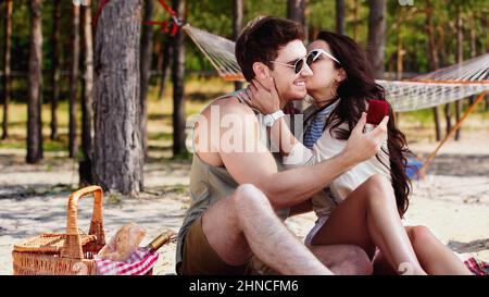 Junge Frau in Sonnenbrille küsst Freund mit Schmuckkachtel in der Nähe Korb mit Wein am Strand Stockfoto