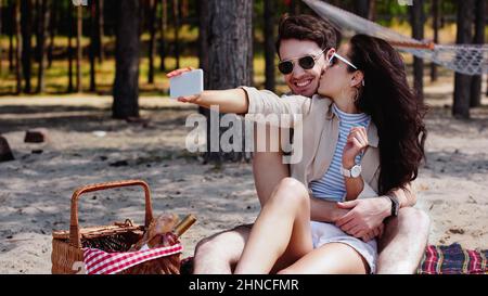 Frau küsst Freund in Sonnenbrillen und macht Selfie beim Picknick am Strand Stockfoto