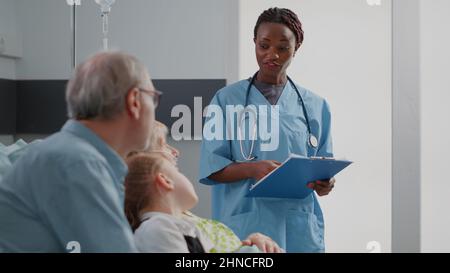 afroamerikanische Krankenschwester im Gespräch mit älteren Patienten und Familie auf der Krankenstation. Medizinische Assistentin erklärt älteren Frauen und Besuchern die Diagnose. Facharzt für Gesundheitsfürsorge diskutiert Behandlung. Stockfoto
