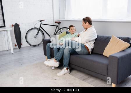 Positives Kind mit Laptop in der Nähe des Vaters auf der Couch Stockfoto