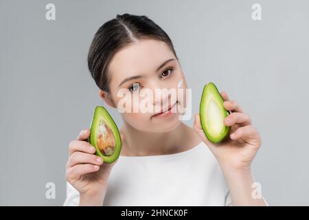 Frau mit Down-Syndrom und Creme auf dem Gesicht mit reifen Avocado isoliert auf grau Stockfoto