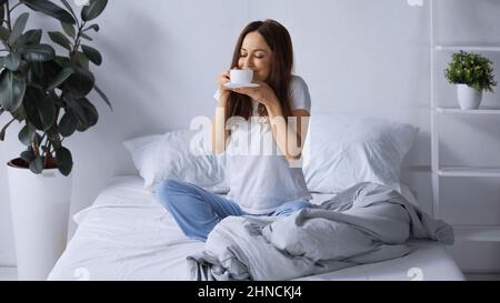 Zufrieden Frau im Schlafanzug sitzt auf dem Bett und riecht aromatischen Kaffee Stockfoto