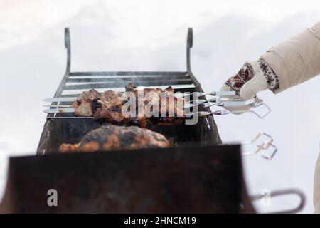 Eine Frauenhand in einem Fäustling fritiert an einem sonnigen Wintertag einen Grill auf dem Grill in der Natur. Selektiver Fokus. Nahaufnahme Stockfoto