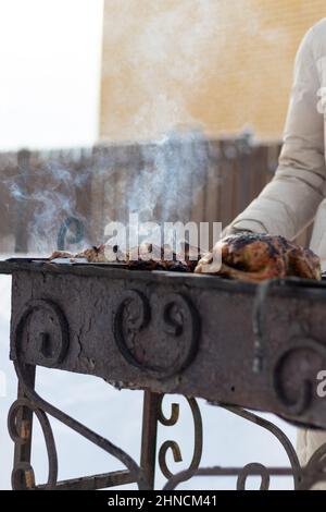 Eine Frauenhand in einem Fäustling fritiert an einem sonnigen Wintertag einen Grill auf dem Grill in der Natur. Selektiver Fokus. Nahaufnahme Stockfoto