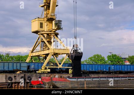 Flusshafenkran mit Clamshell oder Greifer laden Kohle auf Fluss ziehen Boote oder Lastkähne am Pier an bewölktem Tag festgemacht. Energie, Mineralien, Umwelt Stockfoto