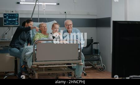 Familie, die Fernsehen schaut und ältere Patienten auf der Krankenhausstation besucht. Ältere Frau, die von Kind und Tochter besucht wird und zur Heilung von Krankheiten ins Krankenhaus eingeliefert wird. Leute, die Spaß mit dem fernsehen haben. Stockfoto