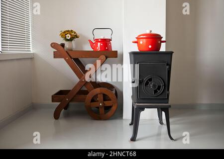 Eisenofen zu Hause Innenraum im Landhaus. Ofen wird in der Nähe des Fensters beheizt. Rotes Geschirr auf dem Tisch Stockfoto