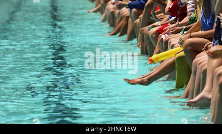 Ein heller, türkisblauer Swimmingpool mit Studenten, die ihre Füße und Zehen im Wasser baumeln lassen, um zuzuschauen. High School Schwimmen Karneval oder Club rac Stockfoto