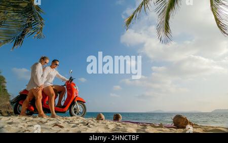 Motorroller Road Trip. Schönes Paar auf rotem Motorrad in weißen Kleidern am Sandstrand. Hochzeit gerade verheiratete Menschen, die in der Nähe von tropischen Palmen, Meer. Flitterwochen am Meer. Asien Thailand. Motorradmiete. Stockfoto
