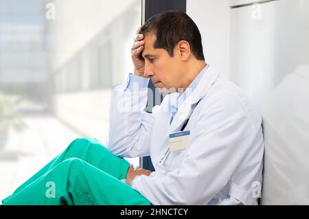 Ein kurzer Schuss eines hübschen jungen Arztes mit Kopfschmerzen, der seinen Kopf hielt, während er sich in ihrem Krankenhaus an eine Wand lehnte Stockfoto