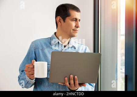 Aufnahme eines hübschen jungen Geschäftsmannes, der Kaffee trinkt, während er in seinem Home-Office an einem Laptop arbeitet und aus dem Fenster blickt Stockfoto
