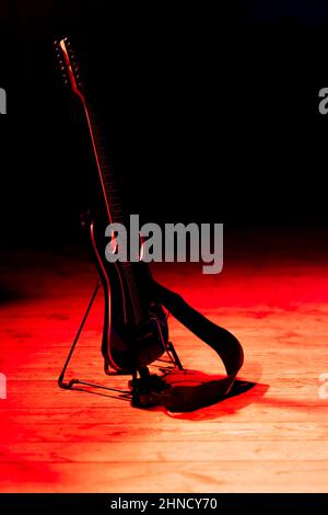 Eine schwarze E-Gitarre, die auf einem auf der Bühne platzierten Ständer basiert. Motiv wird durch starkes rotes Licht beleuchtet. Stockfoto