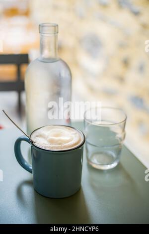 Tasse frisch gebrühten Kaffee mit Schaum serviert mit Löffel auf dem Tisch mit einer Flasche Wasser und Glas auf verschwommenem Hintergrund Stockfoto