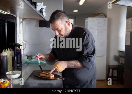 Konzentrierter bärtiger Mann, der eingelegten gesalzenen Fisch auf Toast setzt, der an Bord serviert wird, während er an der Theke in der Küche des Restaurants steht Stockfoto