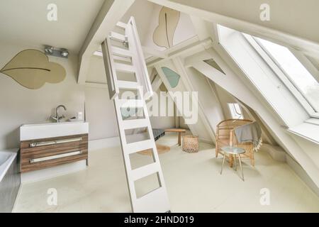 Mansardenzimmer mit Badewanne in der Nähe des Waschbecken im hellen geräumigen Badezimmer mit weißer Leiter in der Nähe verschiedener Dekorationen Stockfoto