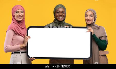 Drei muslimische Frauen posieren mit modernem Smartphone mit leerem Bildschirm über gelbem Studiohintergrund Stockfoto
