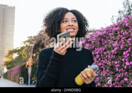 Positive afroamerikanische Frau mit Orangensaft, die aufschaute und Audiobotschaft auf dem Smartphone aufzeichnete, während sie mit purpl in der Nähe von blühenden Büschen stand Stockfoto