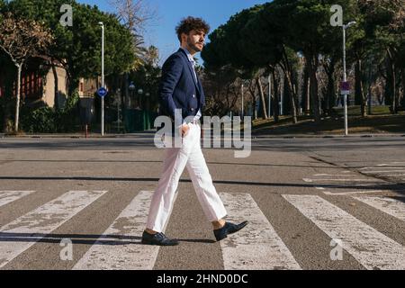 Ganzkörper-Seitenansicht des eleganten hispanischen Mannes, der auf einer Fußgängerüberfahrt auf einer sonnigen Straße mit grünen Bäumen in der Stadt läuft Stockfoto
