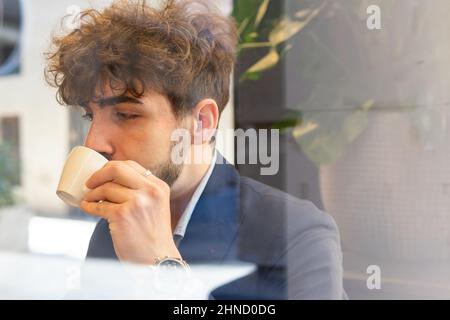 Durch das Fenster des bärtigen hispanischen nachdenklichen männlichen Unternehmers, der auf der Straße der Stadt hinter Glas heißen Kaffee trinkt Stockfoto