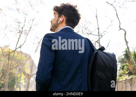 Von der unteren Seite sehen Sie den bärtigen hispanischen Mann in eleganter Kleidung und Rucksack, der Musik in kabellosen Kopfhörern hört, während er auf der sonnigen Straße i läuft Stockfoto