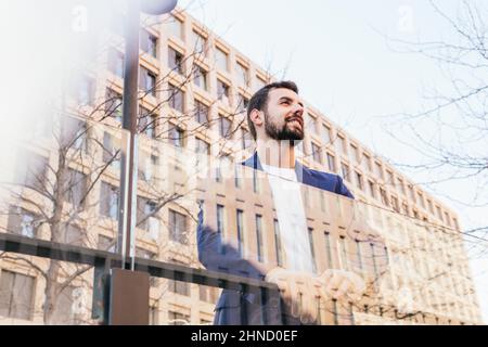 Von unten positive bärtige männliche Unternehmer in elegantem Verschleiß spiegelt sich in einem modernen Gebäude mit Glaswand auf der Straße der Stadt mit Bäumen Stockfoto