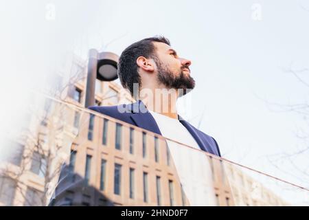 Von unten positive bärtige männliche Unternehmer in elegantem Verschleiß spiegelt sich in einem modernen Gebäude mit Glaswand auf der Straße der Stadt mit Bäumen Stockfoto