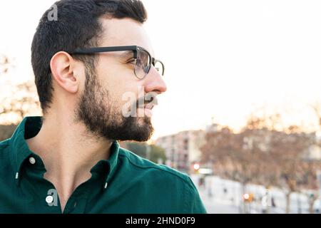 Seriöse bärtige männliche Unternehmerin in eleganter Kleidung und stilvollen Brillen blicken weg, während sie auf der Straße mit Bäumen in der Stadt steht Stockfoto