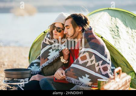 Ein paar Forscher, eingewickelt in warmes Karo, sitzen in der Nähe des Zeltes auf dem Campingplatz und genießen das Wochenende am Seeufer Stockfoto