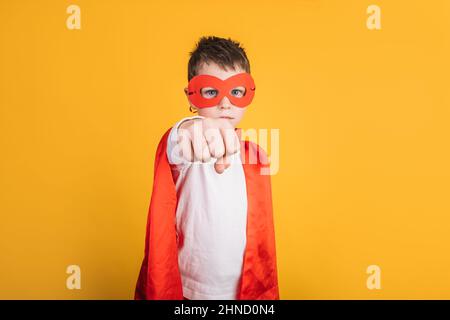 Kind in roter Superhelden-Kleidung und Maske, die den Arm streckt, während es im Studio vor gelbem Hintergrund steht und einen bildgebenden Flug führt Stockfoto