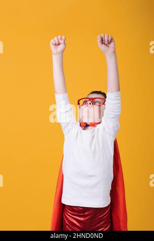 Isolierter Junge in Brille und rotem Superhelden-Kostüm, der aufschaut, während er vorgibt, im Studio auf gelbem Hintergrund zu fliegen Stockfoto