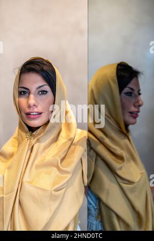 Erwachsene ethnische weibliche Person im eleganten goldenen Kopftuch, die neben dem Spiegel steht und tagsüber zu Hause die Kamera anschaut Stockfoto