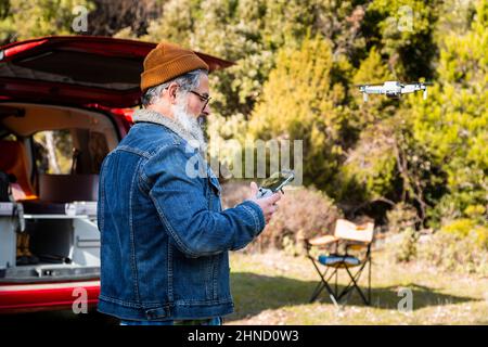 Seitenansicht eines bärtigen, reifen Mannes, der eine moderne fliegende Drohne mit Fernbedienung betreibt, während er in der Nähe eines Mini-Campers auf dem Land steht Stockfoto