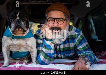 Älterer bärtiger Rüde mit Bart, der auf die Kamera schaut, während er im Mini-Camper mit der französischen Bulldogge auf dem Landweg liegt Stockfoto