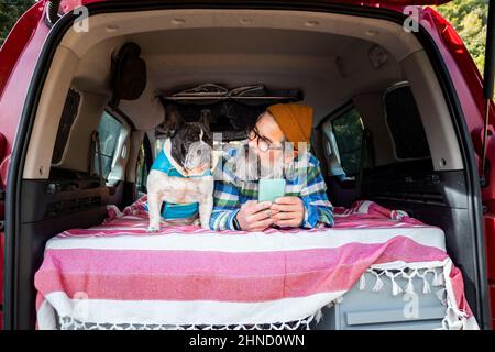 Reifer Mann beim Surfen im Smartphone, während er im Mini-Camper mit der französischen Bulldog auf dem Landweg liegt Stockfoto