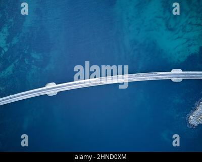 Drohnenansicht einer langen schmalen Brücke über das ruhige klare Meer auf grasbewachsenen Lofoten-Inseln mit Gebäuden in der Natur Norwegens Stockfoto
