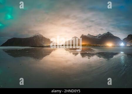 Malerische Landschaft mit ruhigem Meer, die massive felsige Berge mit schneebedeckten Gipfeln während der Ebbe am wolkigen Sonnenuntergang in den Lofoten reflektiert Stockfoto