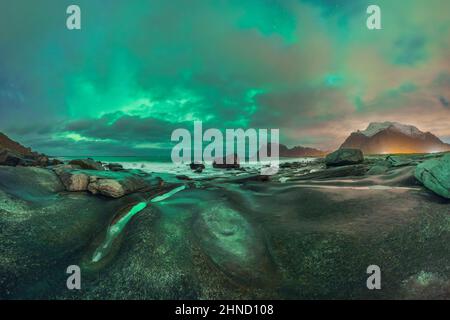 Spektakuläre Landschaft der felsigen Küste des schäumenden Ozeans gegen den bewölkten Abendhimmel mit hellen Polarlichtern auf den Lofoten-Inseln Stockfoto