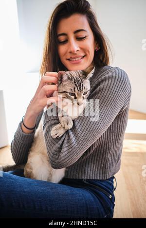Optimistische weibliche Besitzerin streichelt und entzückende Katze, während sie auf dem Boden des hellen Wohnzimmers sitzt Stockfoto