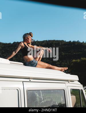 Ganzkörper-Seitenansicht der angenehmen barfuß nachdenklichen Hippie weibliche Reisende sitzen mit geschlossenen Augen auf dem Dach des weißen Camping van in der Natur geparkt d Stockfoto
