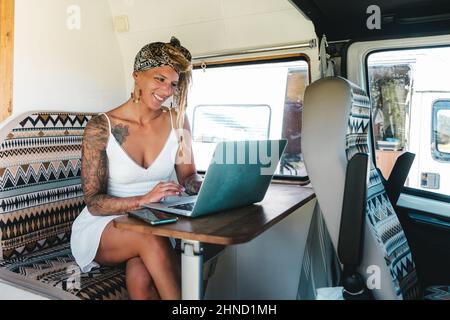 Optimistische Hipster-Hündin in weißem Kleid, die auf einem modernen Netbook tippt, während sie auf der Straße in einem leichten Campingbus neben dem Fenster sitzt Stockfoto
