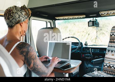 Rückansicht einer optimistischen Frau in weißem Kleid, die auf einem modernen Netbook tippt, während sie während einer Autoreise in einem leichten Campingbus neben dem Fenster sitzt Stockfoto