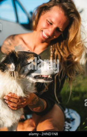 Positive weibliche Besitzerin umarmt niedlichen haarigen australischen Schäferhund mit gefleckten Schnauze, während sie am Sommertag auf der sonnigen Straße sitzt Stockfoto