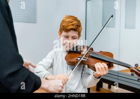 Crop-Musiklehrer in der Nähe von Rotschopf Junge mit Geige im Klassenzimmer stehen Stockfoto